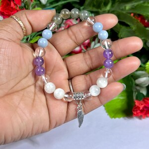 May include: A beaded bracelet featuring a variety of gemstones in shades of clear, purple, blue, and green. The bracelet has a silver charm in the shape of a wing. The bracelet is held in a hand.
