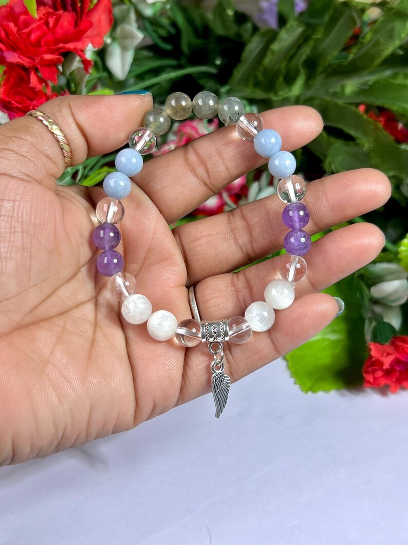 May include: A beaded bracelet featuring a variety of gemstones in shades of clear, purple, blue, and green. The bracelet has a silver charm in the shape of a wing. The bracelet is held in a hand.