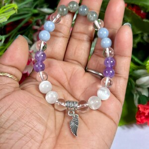 May include: A beaded bracelet featuring a variety of gemstones in shades of purple, blue, clear, and white. The bracelet includes a silver-colored charm in the shape of a wing. The bracelet is strung on a clear elastic cord.