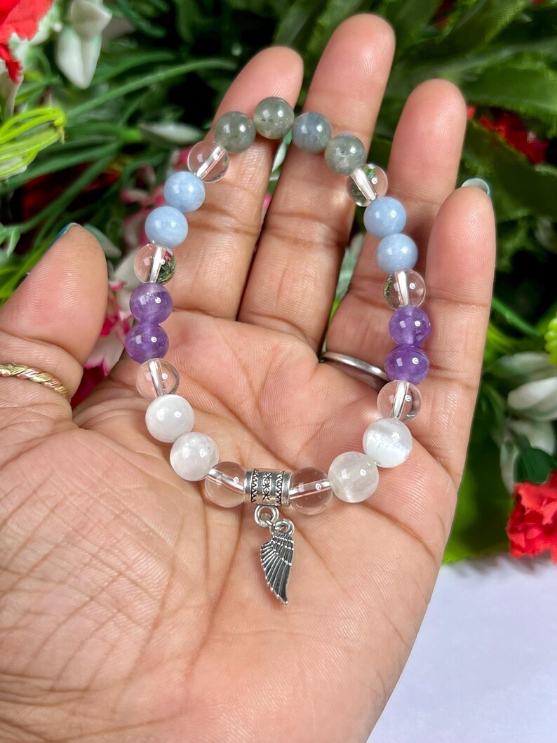 May include: A beaded bracelet featuring a variety of gemstones in shades of purple, blue, clear, and white. The bracelet includes a silver-colored charm in the shape of a wing. The bracelet is strung on a clear elastic cord.