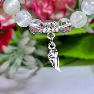 May include: A close-up of a beaded bracelet featuring white and clear beads. A silver charm in the shape of a wing hangs from the bracelet. The background is blurred with pink and green foliage.