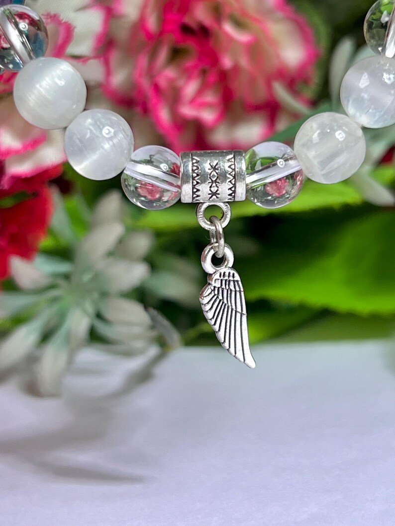 May include: A close-up of a beaded bracelet featuring white and clear beads. A silver charm in the shape of a wing hangs from the bracelet. The background is blurred with pink and green foliage.