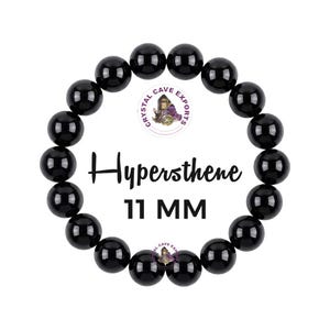 Puede incluir: Una pulsera circular hecha de cuentas de hiperstena negras, lisas y pulidas. El texto Hypersthene 11 MM está impreso en el centro, junto con un pequeño logotipo circular con el texto Crystal Cave Exports y una ilustración de cristales.