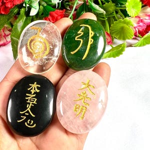 Op de afbeelding: Vier ovale stenen met gouden symbolen in een hand. De stenen zijn helder, groen, zwart en roze. De heldere steen heeft een spiraalsymbool. De groene steen heeft een verticaal symbool. De zwarte steen heeft Japanse karakters. De roze steen heeft Japanse karakters.