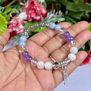 May include: A beaded bracelet featuring a variety of colorful gemstones, including clear, purple, blue, and white beads. The bracelet has a silver charm with a wing design. The bracelet is held in a hand against a backdrop of flowers.