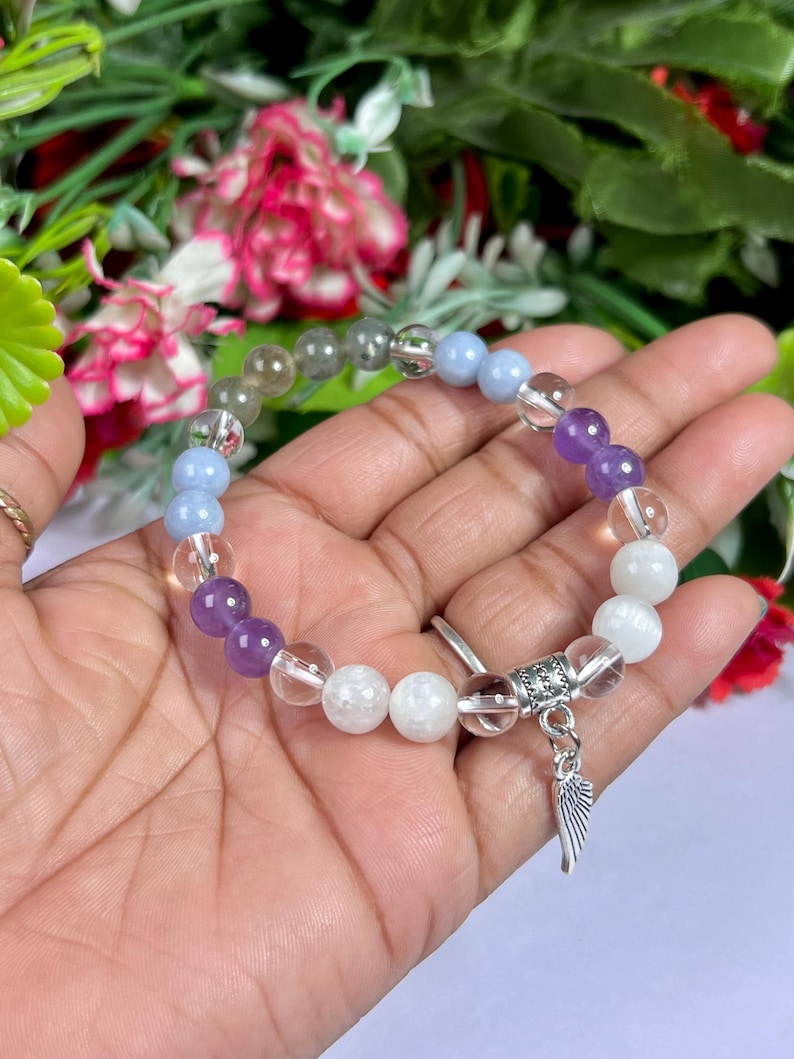 May include: A beaded bracelet featuring a variety of colorful gemstones, including clear, purple, blue, and white beads. The bracelet has a silver charm with a wing design. The bracelet is held in a hand against a backdrop of flowers.