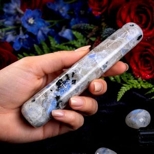 May include: A cylindrical polished stone wand featuring a translucent grey and white base with prominent inclusions of iridescent blue flashes and scattered black mineral flecks throughout the smooth, elongated crystal surface.