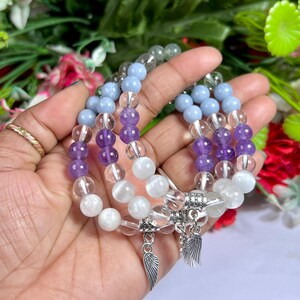 May include: Three beaded bracelets with a variety of gemstone beads in shades of purple, blue, clear, and white. Each bracelet has a silver charm with wing details. The bracelets are displayed against a backdrop of green foliage and flowers.
