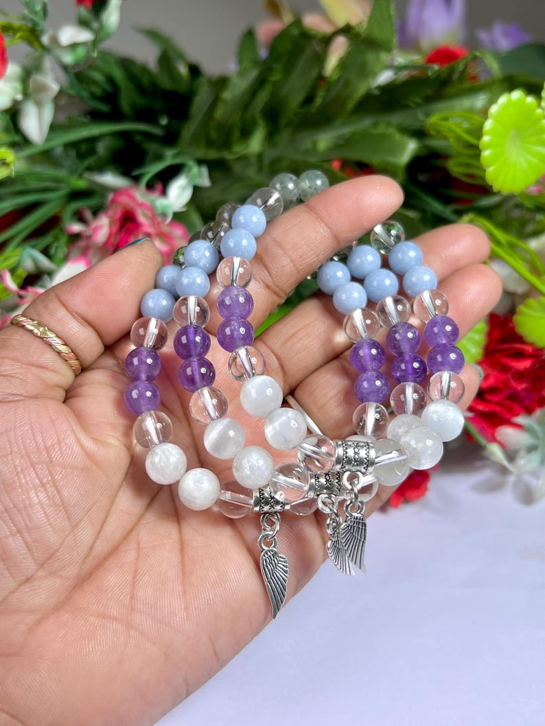 May include: Three beaded bracelets with a variety of gemstone beads in shades of purple, blue, clear, and white. Each bracelet has a silver charm with wing details. The bracelets are displayed against a backdrop of green foliage and flowers.