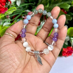 May include: A beaded bracelet featuring a variety of colorful gemstones. The bracelet includes clear, white, light blue, purple, and gray beads. A silver-colored charm in the shape of a wing hangs from the bracelet. The bracelet is approximately 7 inches in circumference.