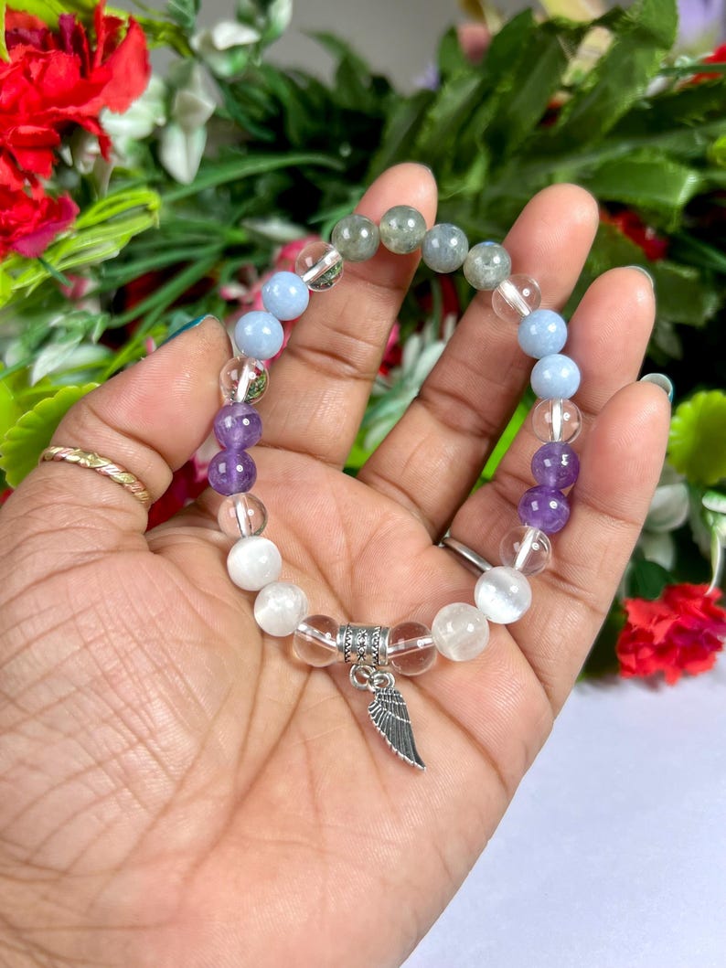 May include: A beaded bracelet featuring a variety of colorful gemstones. The bracelet includes clear, white, light blue, purple, and gray beads. A silver-colored charm in the shape of a wing hangs from the bracelet. The bracelet is approximately 7 inches in circumference.