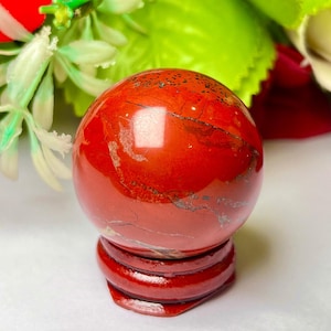 Può includere: Una sfera di diaspro rosso lucido, appoggiata su un supporto in legno abbinato. La sfera presenta motivi naturali e variazioni di colore, con una finitura lucida. Il supporto è rosso scuro, con un design a strati. Lo sfondo include vegetazione e un fiore rosso.