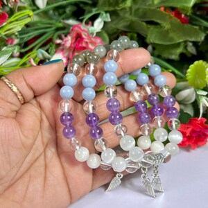 May include: Several beaded bracelets featuring a variety of gemstones, including clear quartz, amethyst, and light blue beads. Each bracelet has a silver charm with a wing design. The bracelets are displayed against a backdrop of colorful flowers.
