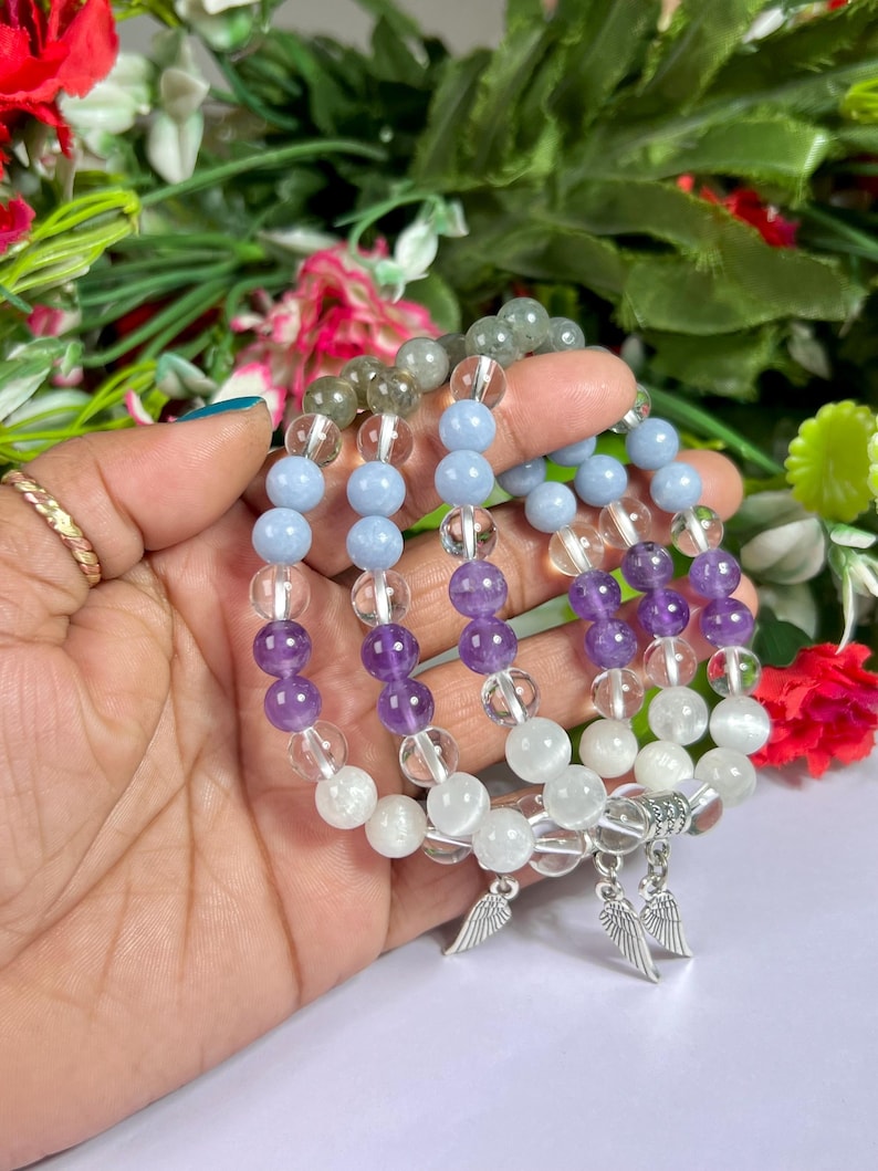 May include: Several beaded bracelets featuring a variety of gemstones, including clear quartz, amethyst, and light blue beads. Each bracelet has a silver charm with a wing design. The bracelets are displayed against a backdrop of colorful flowers.
