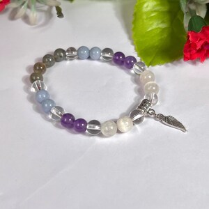 May include: A beaded bracelet featuring a variety of colorful gemstones, including purple, clear, gray, and white beads. The bracelet is accented with a silver-colored charm in the shape of a wing. The bracelet is displayed on a white surface.