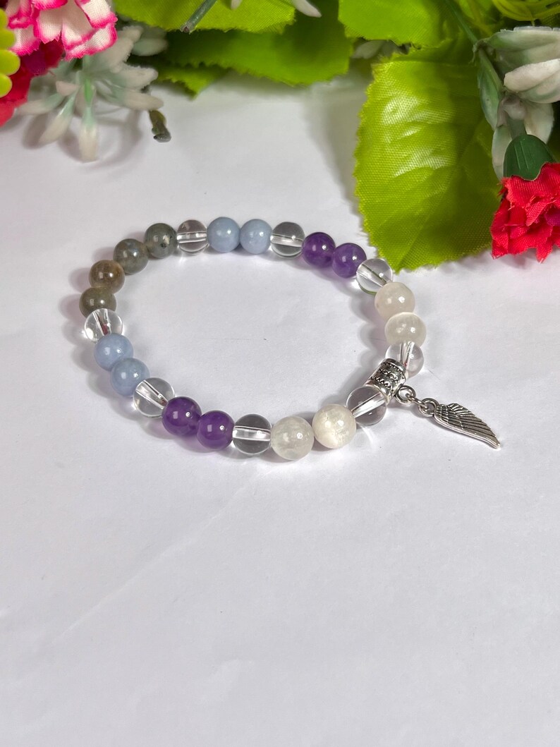May include: A beaded bracelet featuring a variety of colorful gemstones, including purple, clear, gray, and white beads. The bracelet is accented with a silver-colored charm in the shape of a wing. The bracelet is displayed on a white surface.