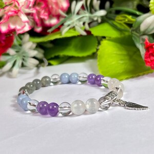 May include: A beaded bracelet featuring a mix of clear, purple, gray, and blue beads. The bracelet has a silver charm in the shape of a feather. The beads are strung on a stretchy cord.