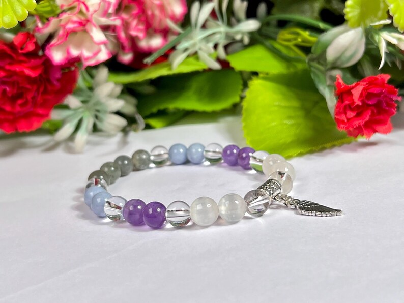 May include: A beaded bracelet featuring a mix of clear, purple, gray, and blue beads. The bracelet has a silver charm in the shape of a feather. The beads are strung on a stretchy cord.