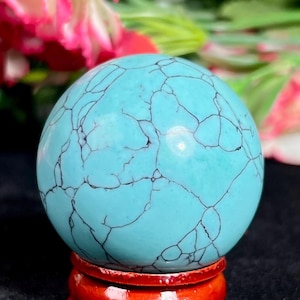 May include: A polished, spherical turquoise stone featuring a vibrant cyan blue colour with intricate, dark black veining patterns throughout its surface. The sphere is securely positioned on a small, circular, glossy reddish-brown wooden stand.