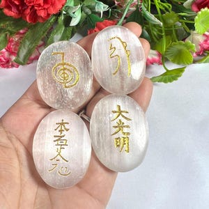 Puede incluir: Cuatro piedras ovaladas de selenita transparente con símbolos y caracteres dorados. Las piedras se sostienen en una mano, con follaje rojo y verde en el fondo. Los símbolos y caracteres son dorados.
