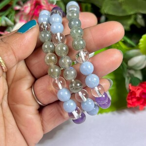 May include: Three beaded bracelets featuring a variety of gemstones. The bracelets include light blue, clear, green, and purple beads. The bracelets are held in a hand with a teal painted fingernail and a gold ring.