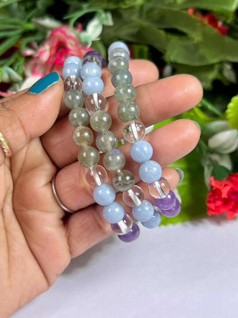 May include: Three beaded bracelets featuring a variety of gemstones. The bracelets include light blue, clear, green, and purple beads. The bracelets are held in a hand with a teal painted fingernail and a gold ring.