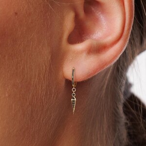 Puede incluir: Primer plano de la oreja de una persona con un pendiente de oro que presenta un pequeño amuleto puntiagudo que cuelga. El pendiente está hecho de una cadena fina con pequeñas cuentas negras.