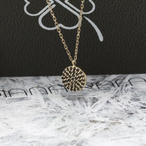 May include: A gold-tone necklace featuring a circular pendant encrusted with small, black gemstones. The fine chain complements the pendant. The backdrop presents a blend of black and white patterns.