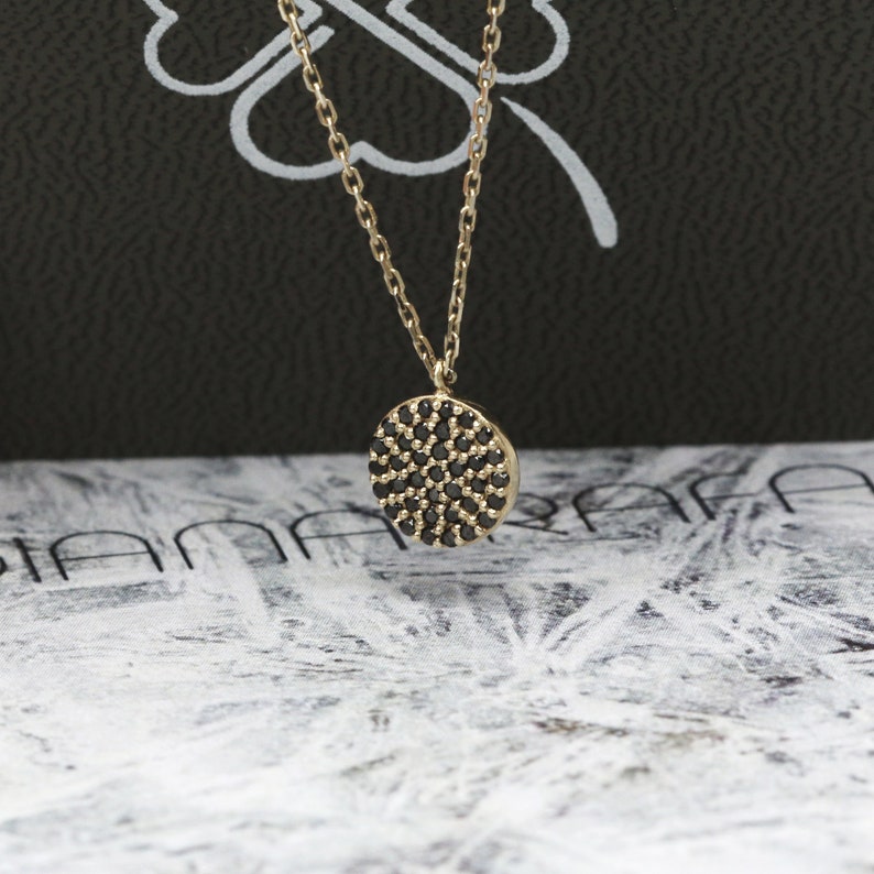 May include: A gold-tone necklace featuring a circular pendant encrusted with small, black gemstones. The fine chain complements the pendant. The backdrop presents a blend of black and white patterns.