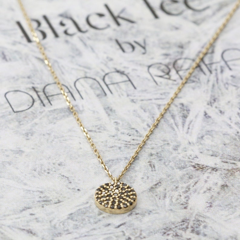 May include: A gold necklace featuring a circular pendant adorned with small black and white stones. The fine chain supports the pendant, set against a patterned backdrop. The text "Black Ice by DIANA RAFA" is visible in the background, adding a branded element.