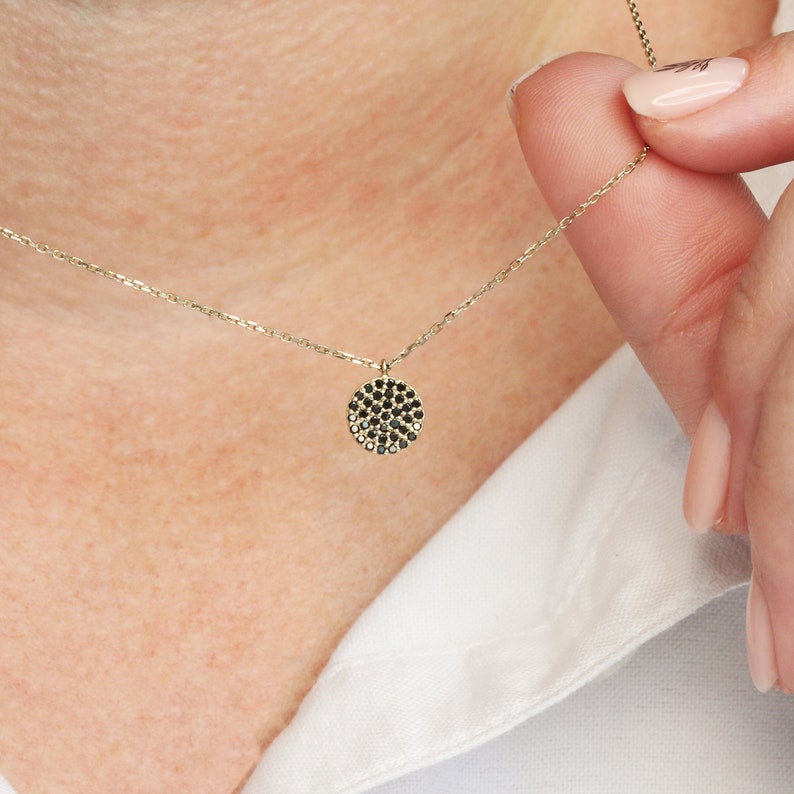 May include: A gold necklace with a small, circular pendant. The pendant is set with a pattern of black and clear gemstones. The necklace is shown against a white shirt.