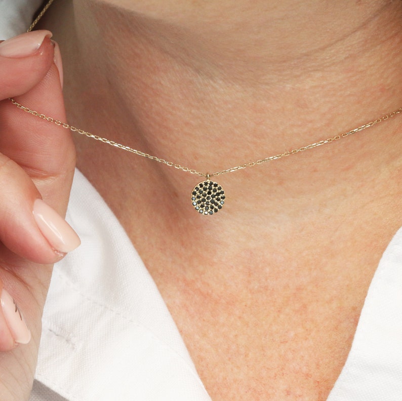 May include: A fine gold necklace with a small, circular pendant. The pendant is set with numerous small, black gemstones, giving a clustered, textured look. The necklace is shown against a white shirt.