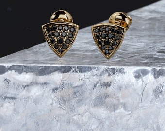 May include: A pair of gold triangle earrings with black diamond accents. The earrings are set on a white, textured surface.
