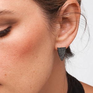 May include: Close-up of a person's ear with a black stud earring. The earring is a triangular shape, covered in small, dark stones. The earring is set against the person's skin and ear.