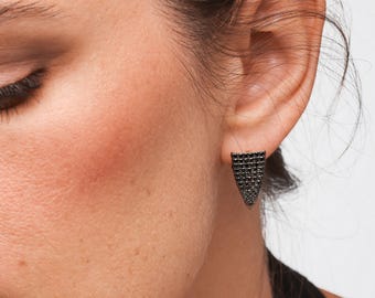 May include: Close-up of a person's ear with a black stud earring. The earring is a triangular shape, covered in small, dark stones. The earring is set against the person's skin and ear.