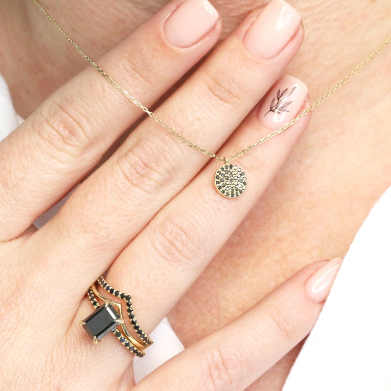 May include: A gold necklace featuring a small, circular pendant set with black gemstones. A gold ring with a large, rectangular black gemstone and smaller black stones is also shown. The jewellery is displayed on a hand.