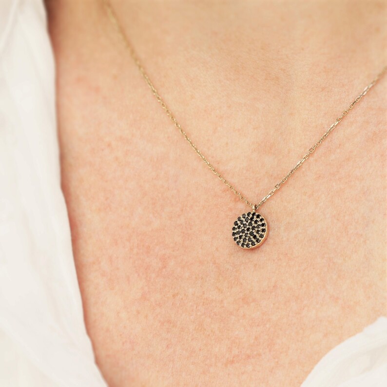 May include: A gold necklace featuring a circular pendant adorned with numerous small, black gemstones. The necklace is displayed against a white shirt.