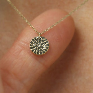 May include: A gold necklace featuring a circular pendant adorned with numerous small, black gemstones. The delicate chain and pendant are displayed, suggesting it is being worn.