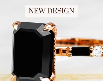 May include: A rose gold engagement ring with a large black emerald cut gemstone and smaller black gemstones on the band. The ring is set on a white marble surface. NEW DESIGN.