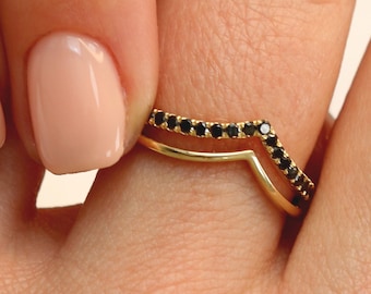 May include: Two gold rings are shown, one a simple V-shape and the other a curved band with small black stones. The rings are on a finger with a light pink nail polish. The rings are likely for fashion or as a wedding band.