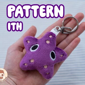 May include: Purple plush starfish keyring with black eyes and white accents. The keyring is held in a hand and has a silver metal ring.