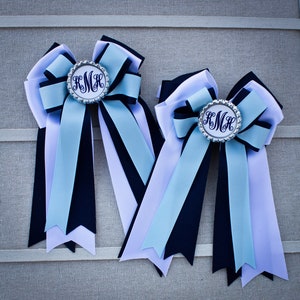 May include: Two light blue, white, and black ribbon horse hair bows with silver monogrammed centerpieces. The monogram reads "K.M.H."