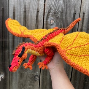 May include: A crocheted dragon toy made with orange, red and brown yarn. The dragon has its wings outstretched and is being held by a hand.