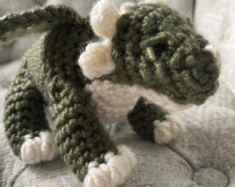 May include: A handmade crochet dragon in shades of green and white. The dragon has a textured appearance with detailed wings, horns, and feet. The dragon is sitting on a light blue textured surface.
