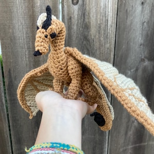 May include: A crocheted dragon with tan and cream-colored wings and body. The dragon has black accents on its horns, nose, and feet. The wings are spread, and the dragon appears to be in flight. The dragon is handmade.