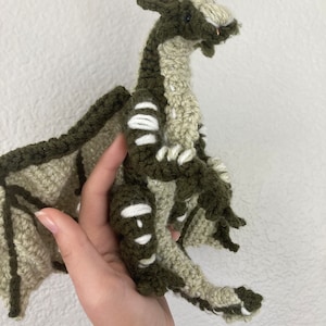 May include: A crocheted green and white dragon with a long tail and wings. The dragon is sitting on a hand.