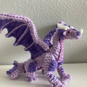 May include: A crocheted purple dragon with white horns and wings. The dragon is standing on a white surface.
