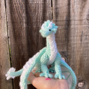 Könnte beinhalten: Ein gehäkelter grüner und weißer Drache mit Flügeln, der auf einer Hand steht.