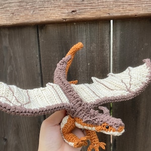 May include: A crocheted dragon toy with brown and white wings, orange legs, and a brown head with black eyes. The dragon is being held in a person's hand.