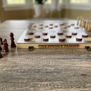 K&ouml;nnte beinhalten: Ein Schach- und Dame-Kombinationsspiel aus Holz mit dunkelbraunen und hellen Holzfiguren. Auf dem Spiel steht der Text "THE SPENCE FAMILY". Das Spiel steht auf einem Holztisch.